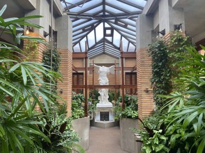 A white marble statue stands at the end of a hallway with brick columns, glass ceiling, and lush green plants on both sides.