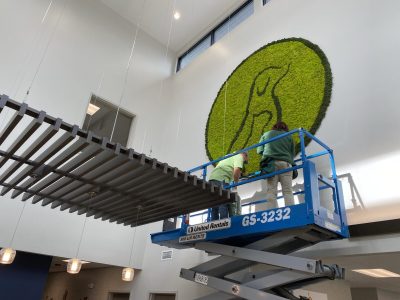 Two people on a scissor lift install a large moss wall art piece featuring a penguin outline in a modern interior.